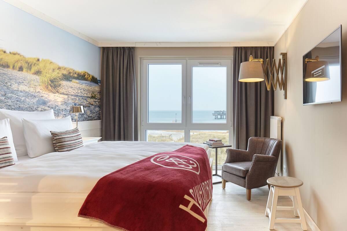 A hotel room with a large bed, a chair, a small stool, a TV, and a view of the beach through the window.