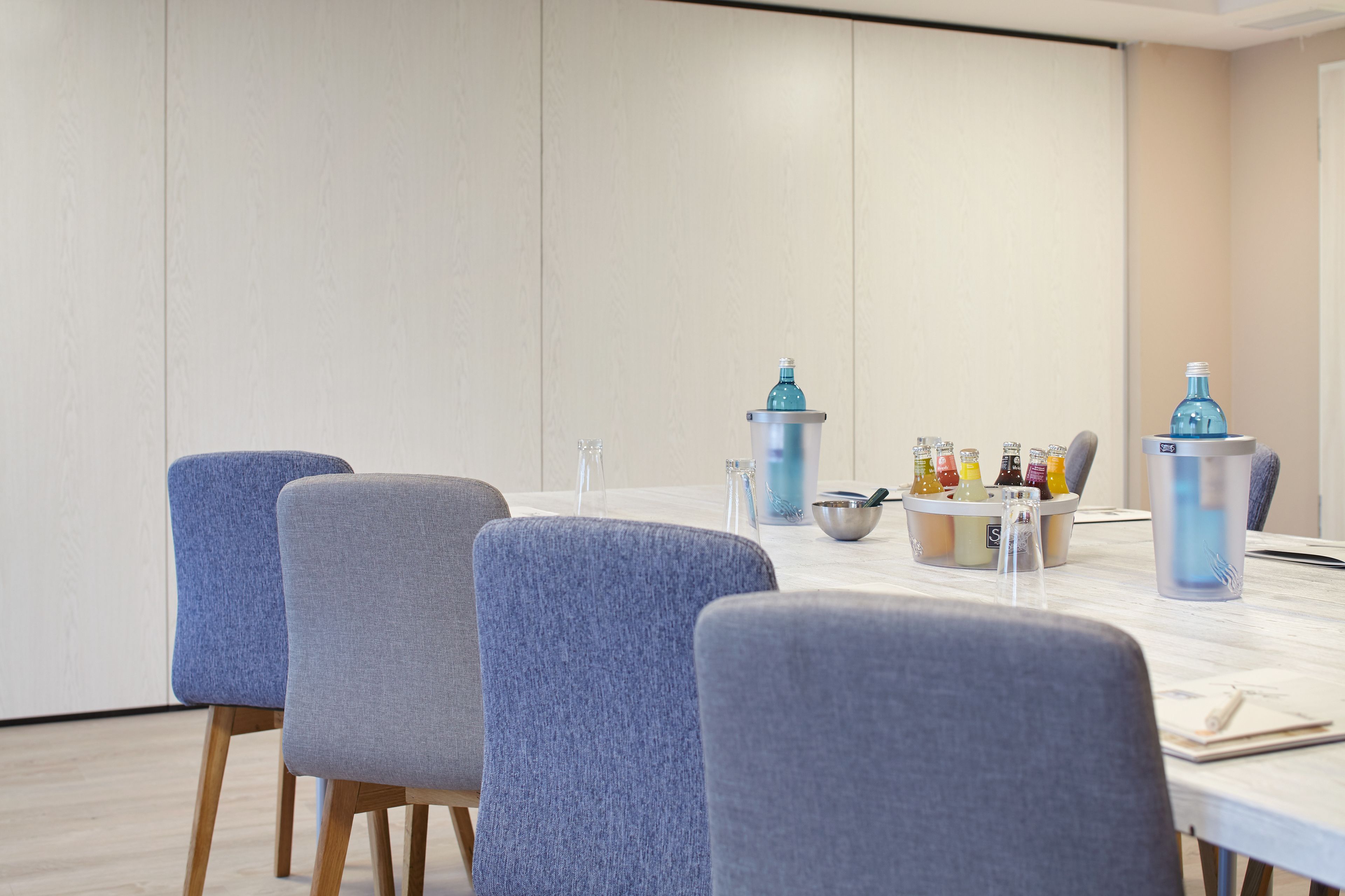 Een vergaderruimte met een lange tafel, vier grijze bekleden stoelen en een waterdispenser.