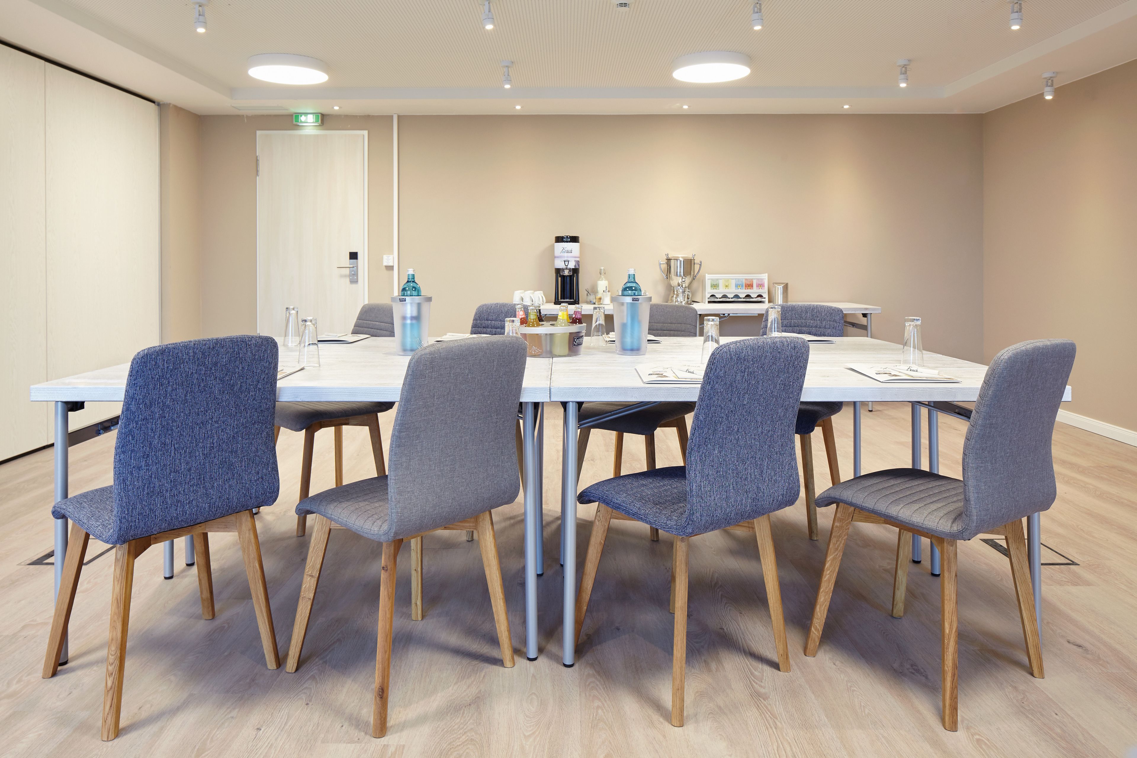 A conference room with a white table, grey upholstered chairs, wooden legs, and a coffee machine.