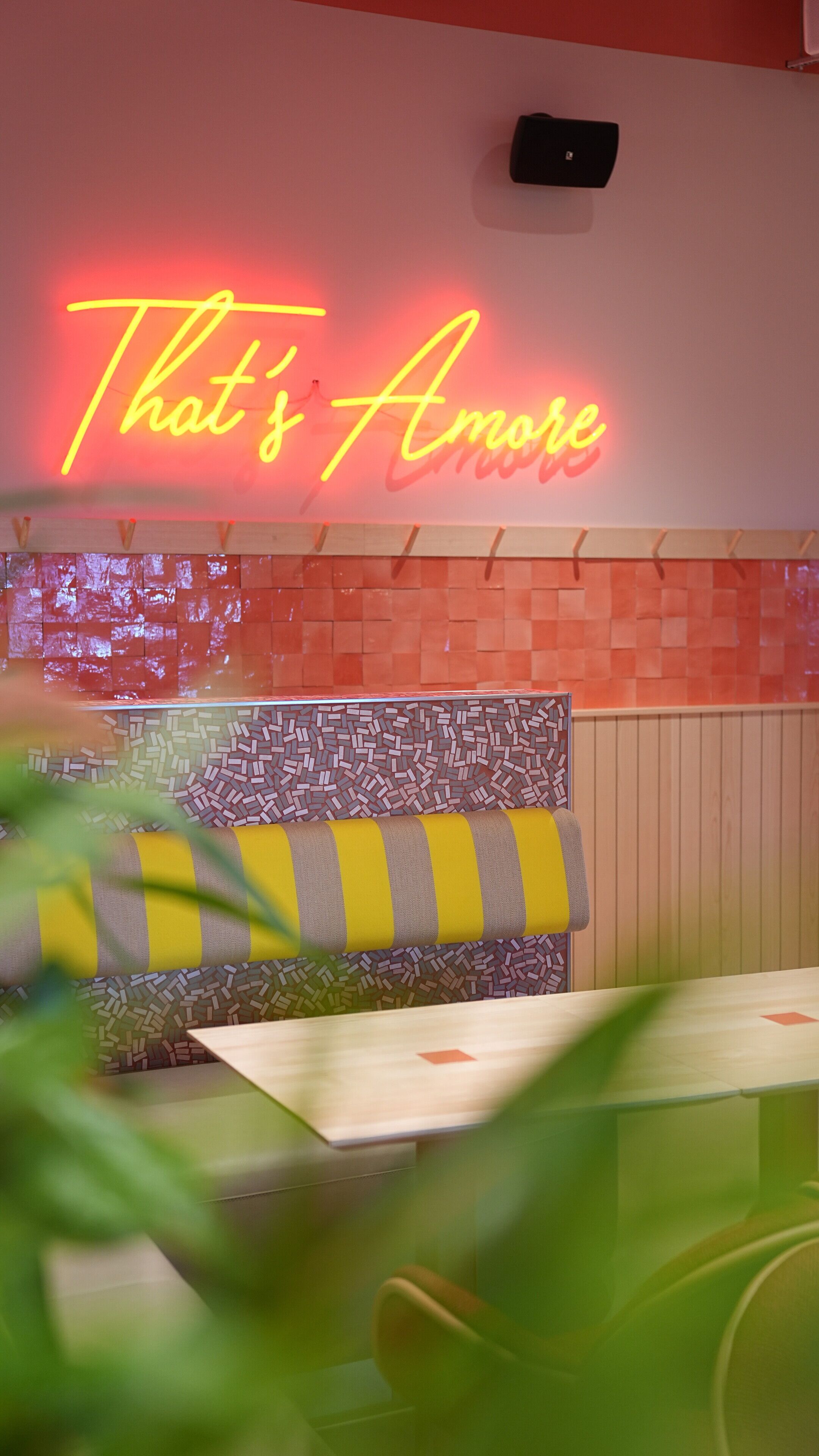 A restaurant interior with a neon sign reading "That's Amore," a patterned wall, and a striped cushion on a bench.