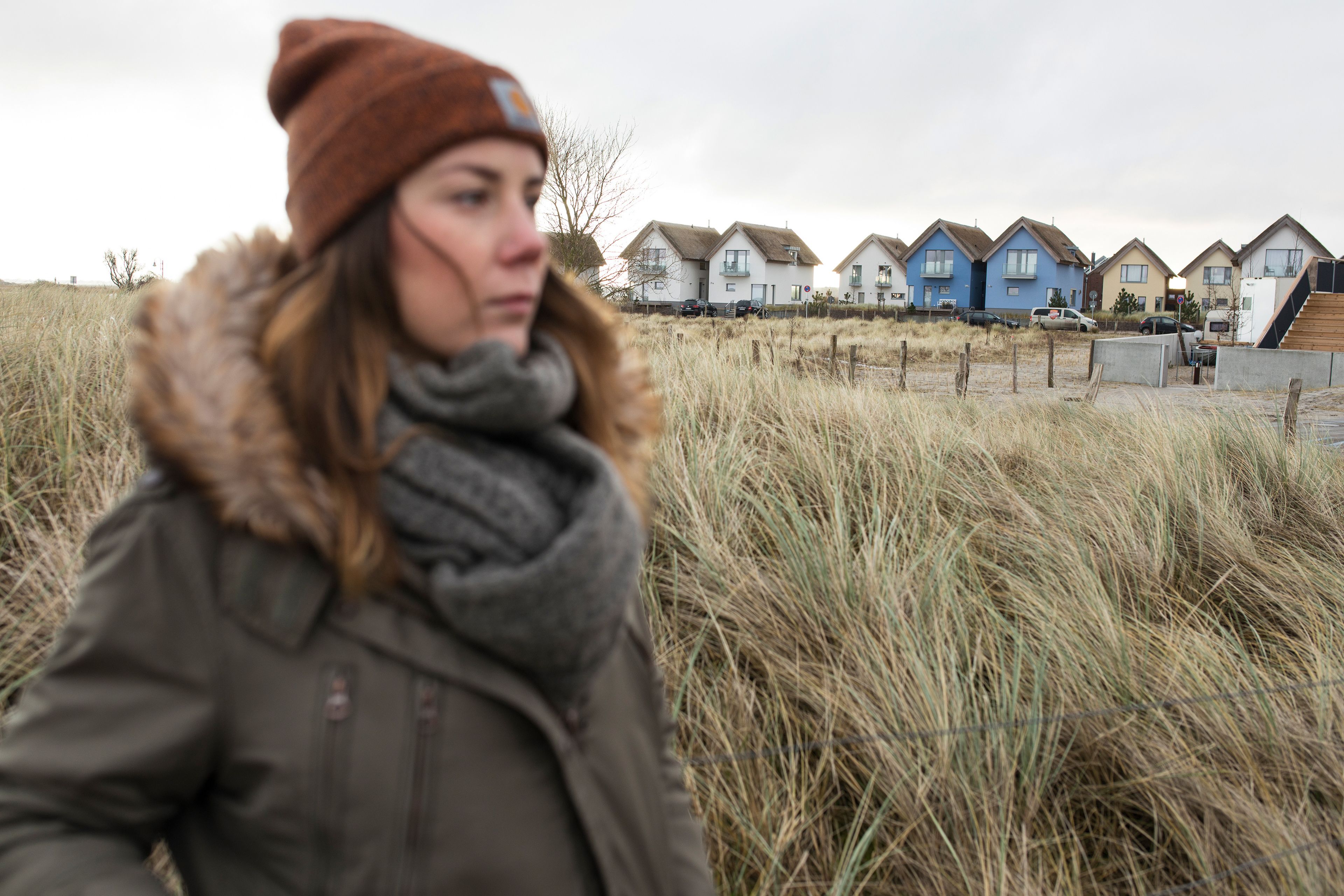 Eine Person mit Jacke und Mütze geht durch ein Feld mit hohem Gras. Im Hintergrund sind mehrere Häuser in verschiedenen Farben zu sehen und eine Holztreppe führt zu einem Gebäude.