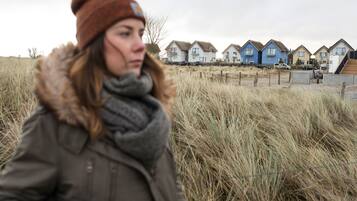 Eine Person mit Jacke und Mütze geht durch ein Feld mit hohem Gras. Im Hintergrund sind mehrere Häuser in verschiedenen Farben zu sehen und eine Holztreppe führt zu einem Gebäude.