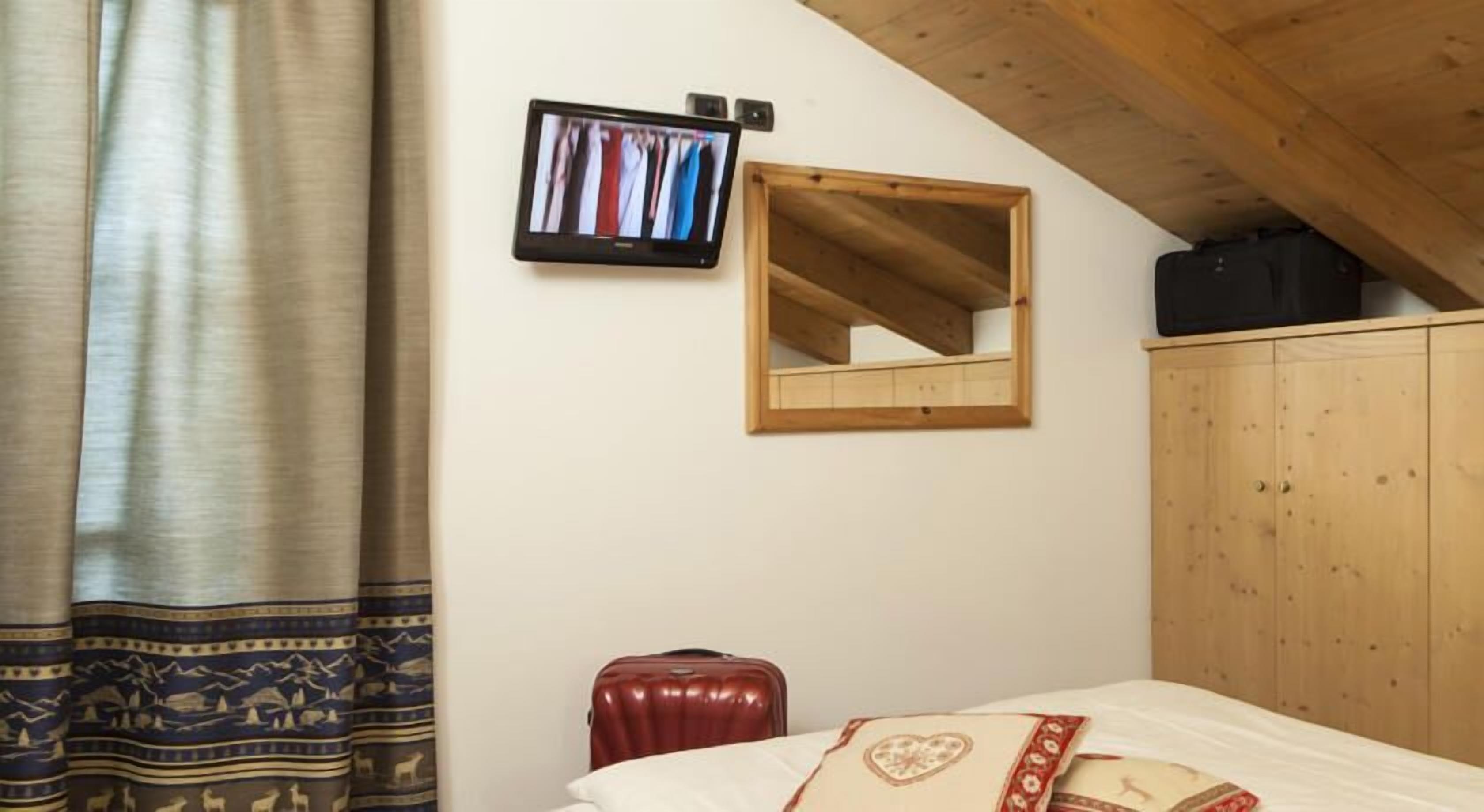 A bedroom with a bed, a wooden wardrobe, a mirror, a television, and a suitcase.
