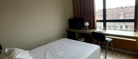 A hotel room with a bed, a desk, a chair, a TV, and a window with a view of buildings.