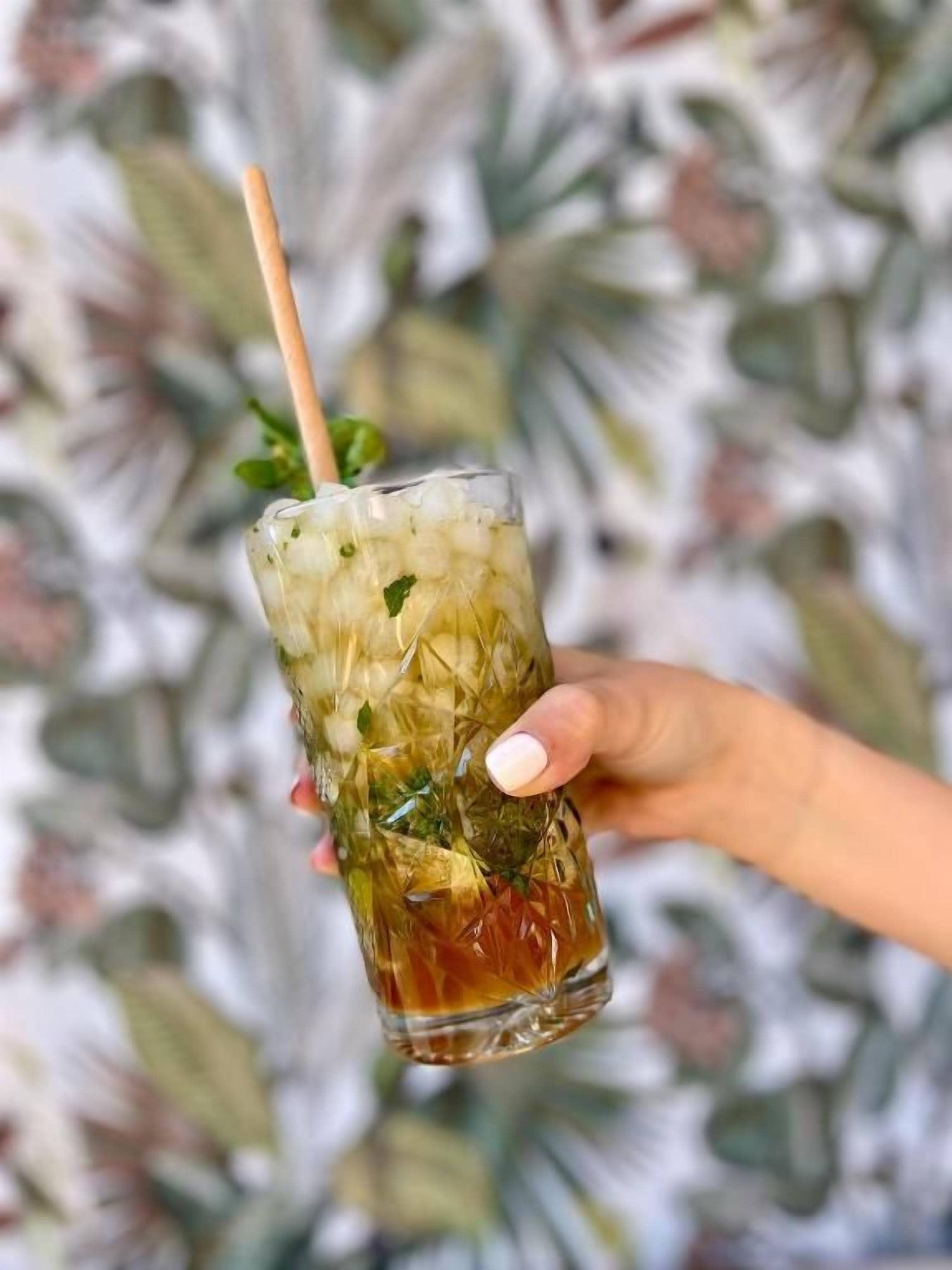 Una mano sosteniendo un vaso con pajita, que contiene una bebida con cubitos de hielo y hojas de menta.