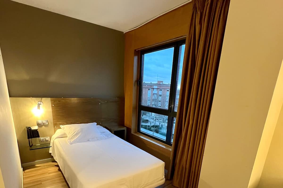 Une chambre d’hôtel avec un lit, un bureau, une chaise, une fenêtre donnant sur des immeubles et un téléphone sur la table de chevet.