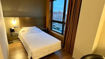 A hotel room with a bed, a desk, a chair, a window with a view of buildings, and a phone on the nightstand.