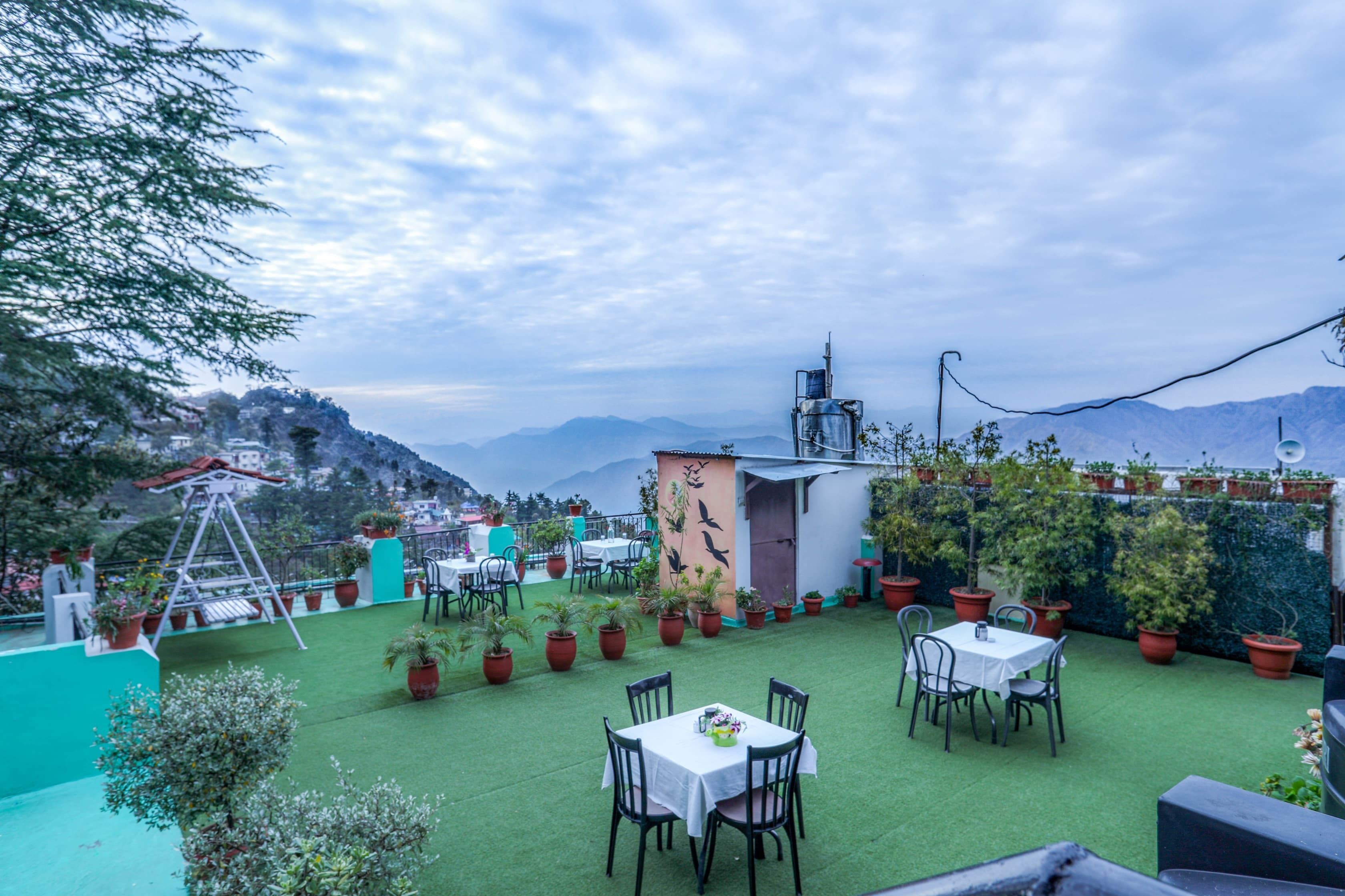 An outdoor patio area with tables and chairs, surrounded by potted plants and a view of mountains.