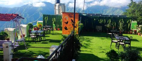 Terraza con mesas y sillas, rodeada de plantas en macetas y vistas a montañas.
