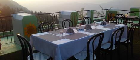 Un área de comedor al aire libre con mesas dispuestas para dos, sillas y vista a las montañas al fondo.