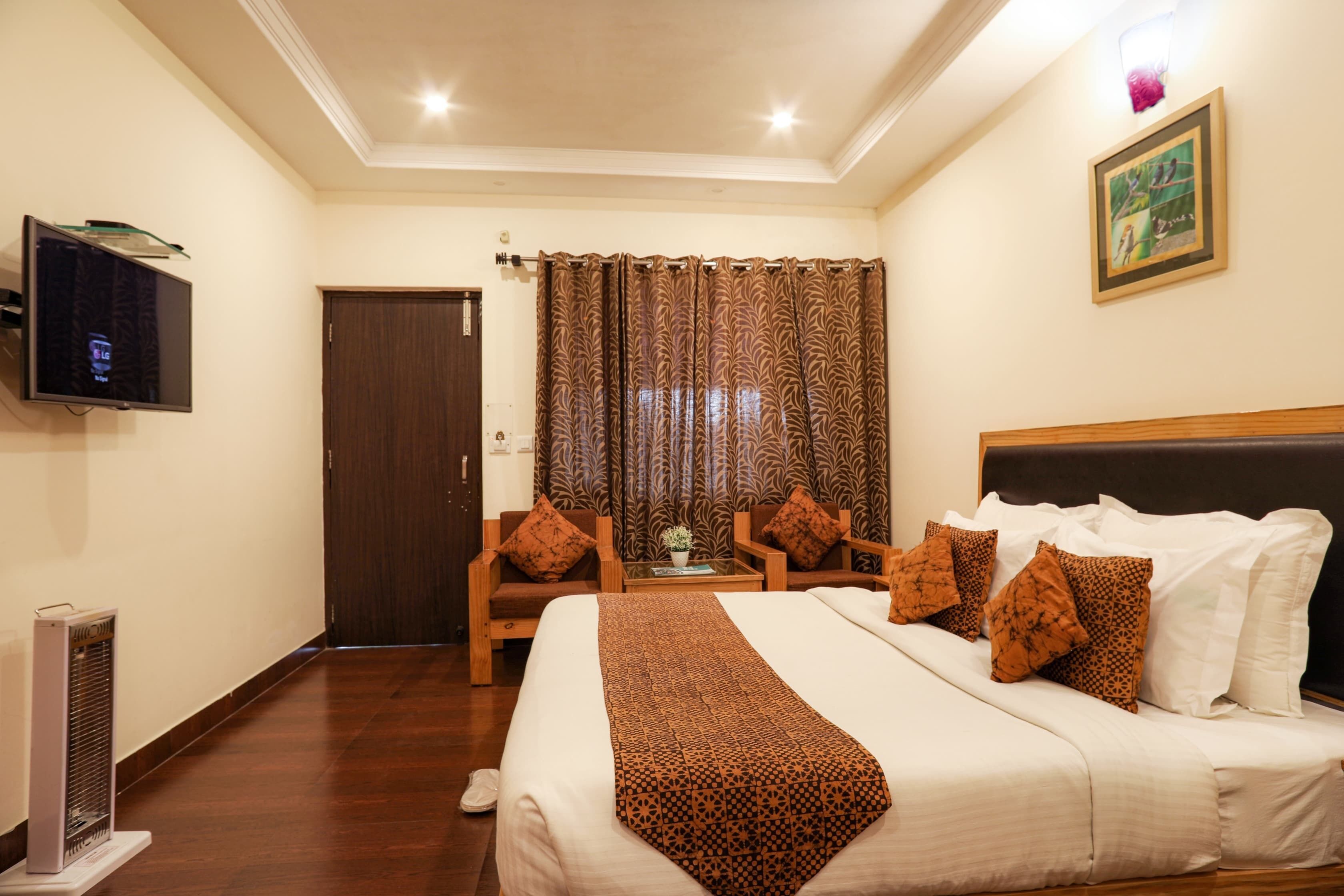 A hotel room with a large bed, two armchairs, a small table with a plant, a television on the wall, and a door leading to another room.