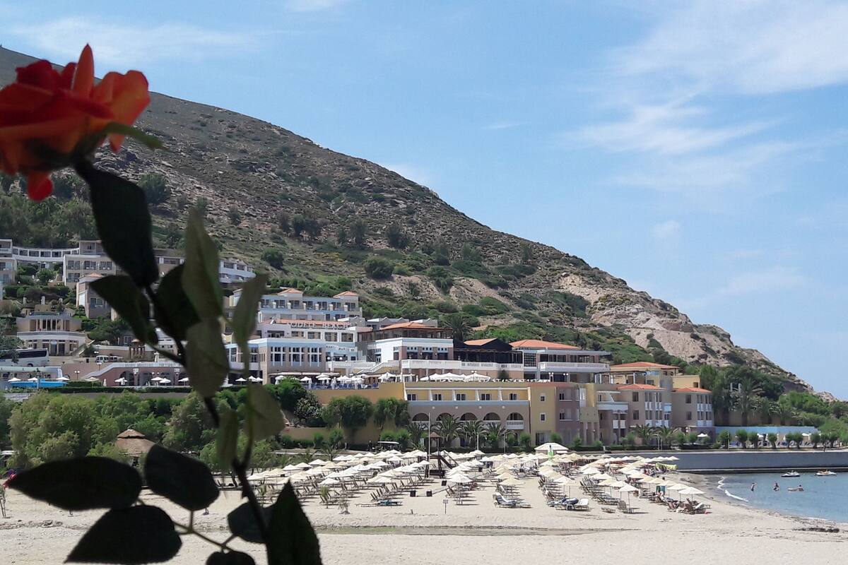 Une plage avec des transats et des parasols, une colline parsemée de bâtiments et un ciel dégagé.