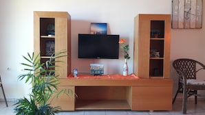 A wooden entertainment center with a flat-screen TV, shelves with decorative items, and a vase with a flower. - Orange Fodele (Malevizi)