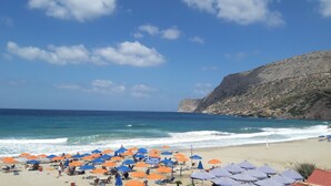 A beach with numerous umbrellas and sun loungers, a rocky cliff in the background, and a clear blue sky. - Orange Fodele (Malevizi)