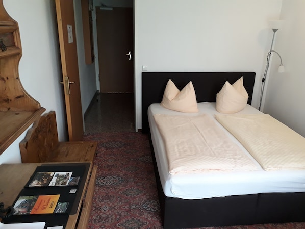 A hotel room with a bed, a wooden dresser, a small table with brochures, and a patterned carpet.