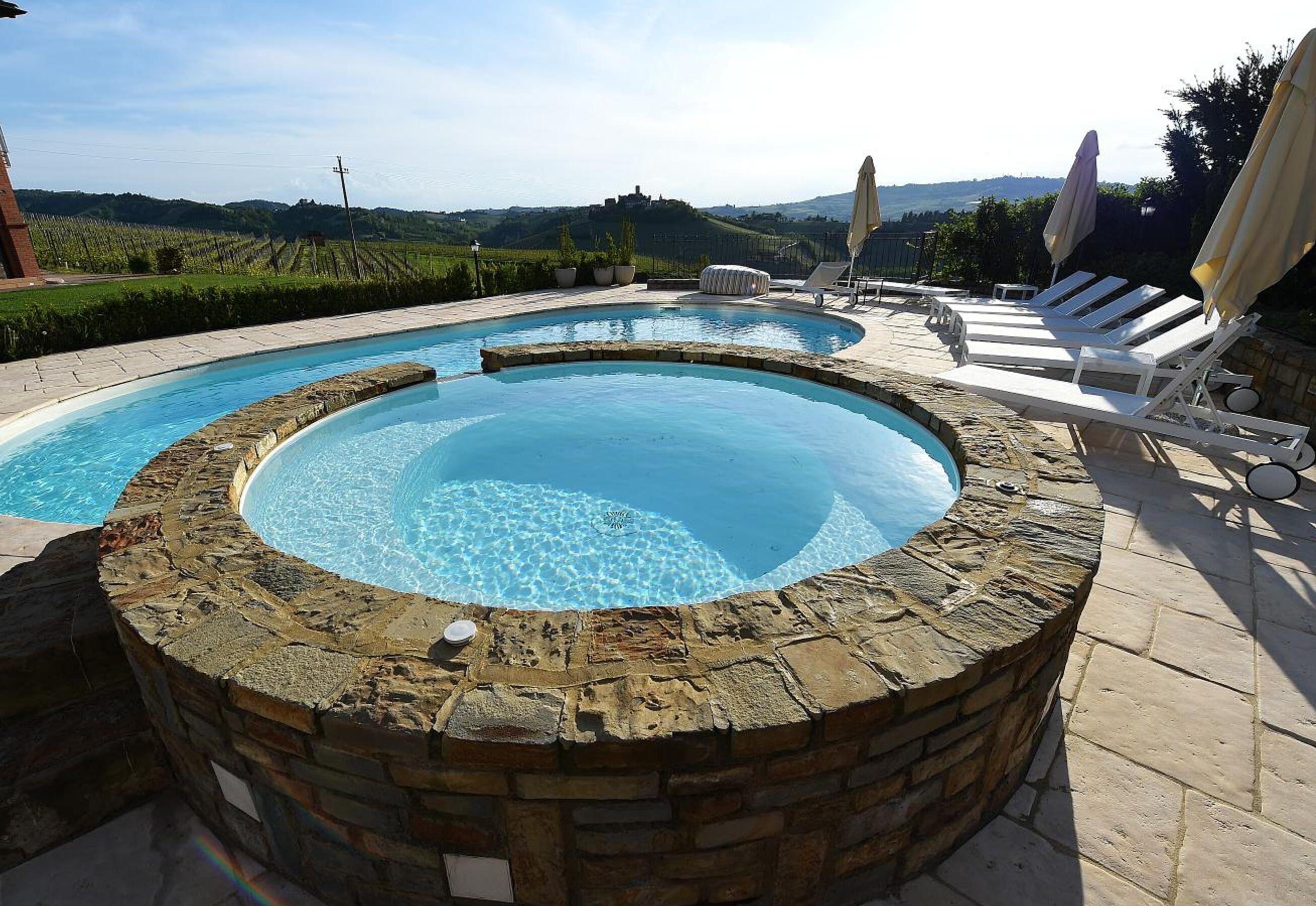 Ein runder Whirlpool mit Steinrand, umgeben von Liegestühlen und Sonnenschirmen, mit Blick auf eine Weinlandschaft.