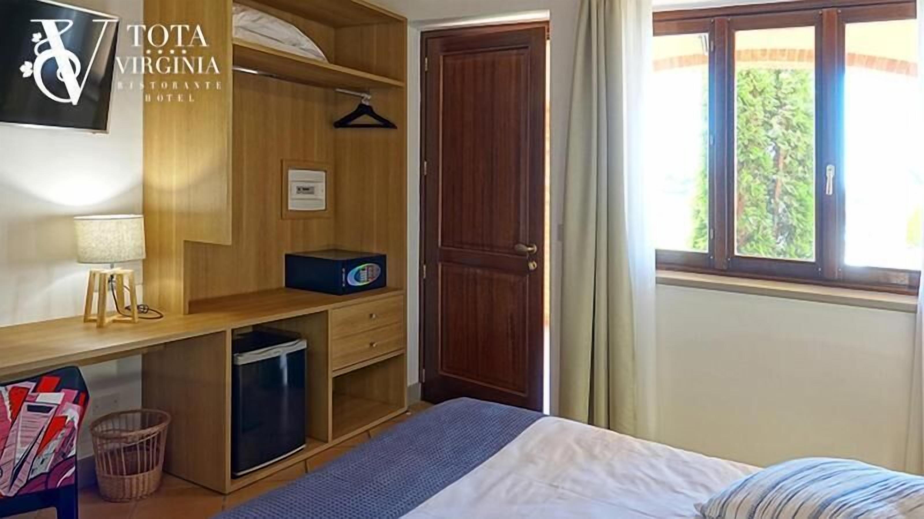 A bedroom with a bed, a desk with a lamp, a wardrobe, a small refrigerator, and a window with a view of greenery.