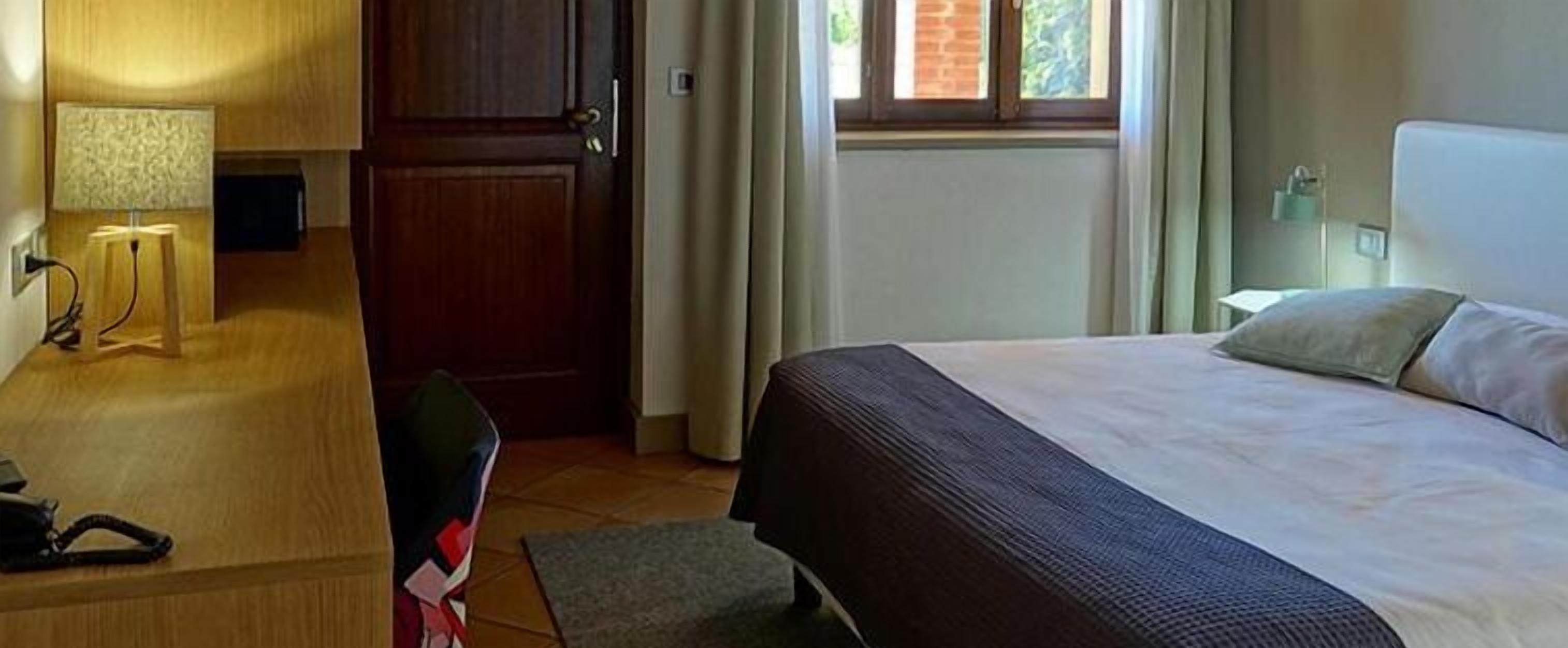 Quarto de hotel com uma cama, escrivaninha com abajur, uma cadeira e janela com cortinas.