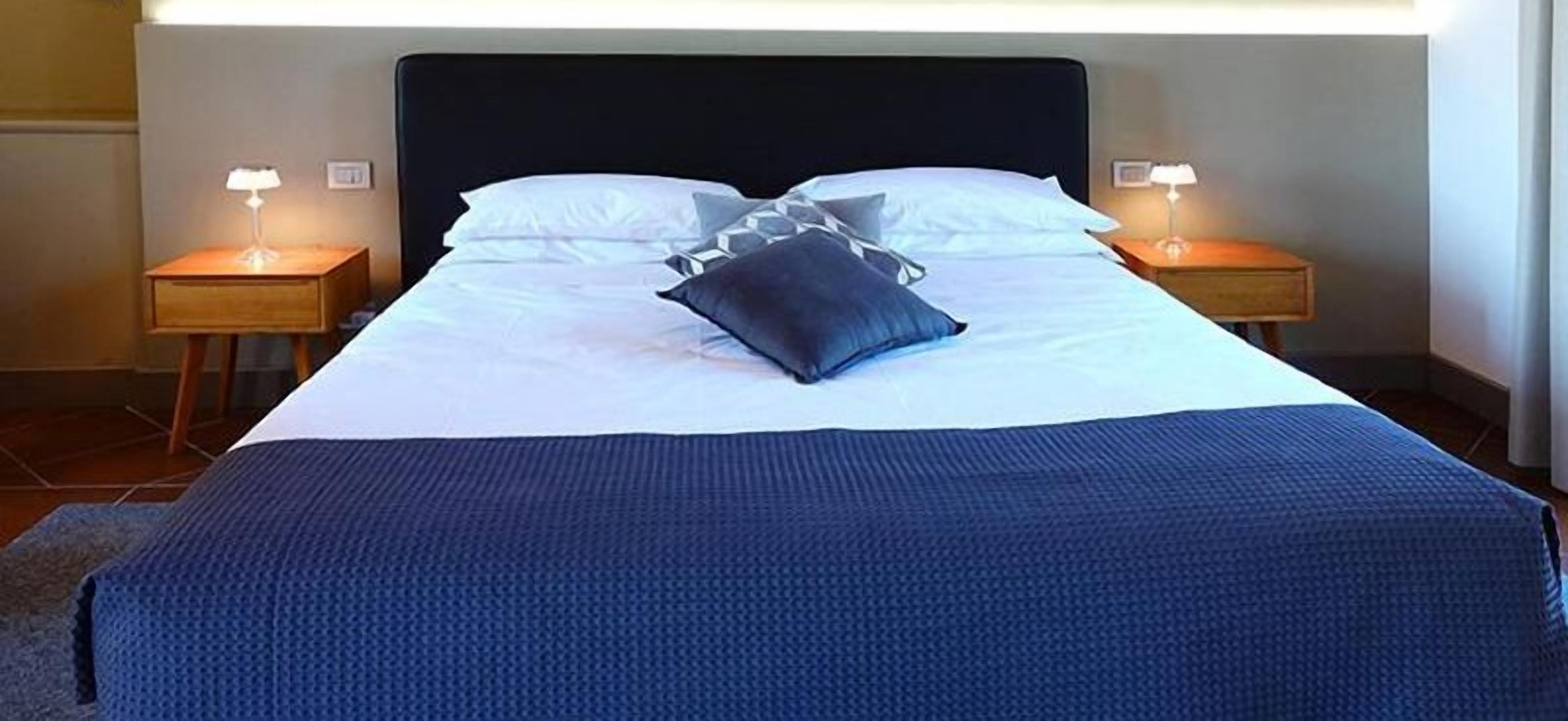 Una cama bien hecha con una manta azul oscuro y almohadas, dos mesitas de noche con lámparas y un cabecero oscuro.