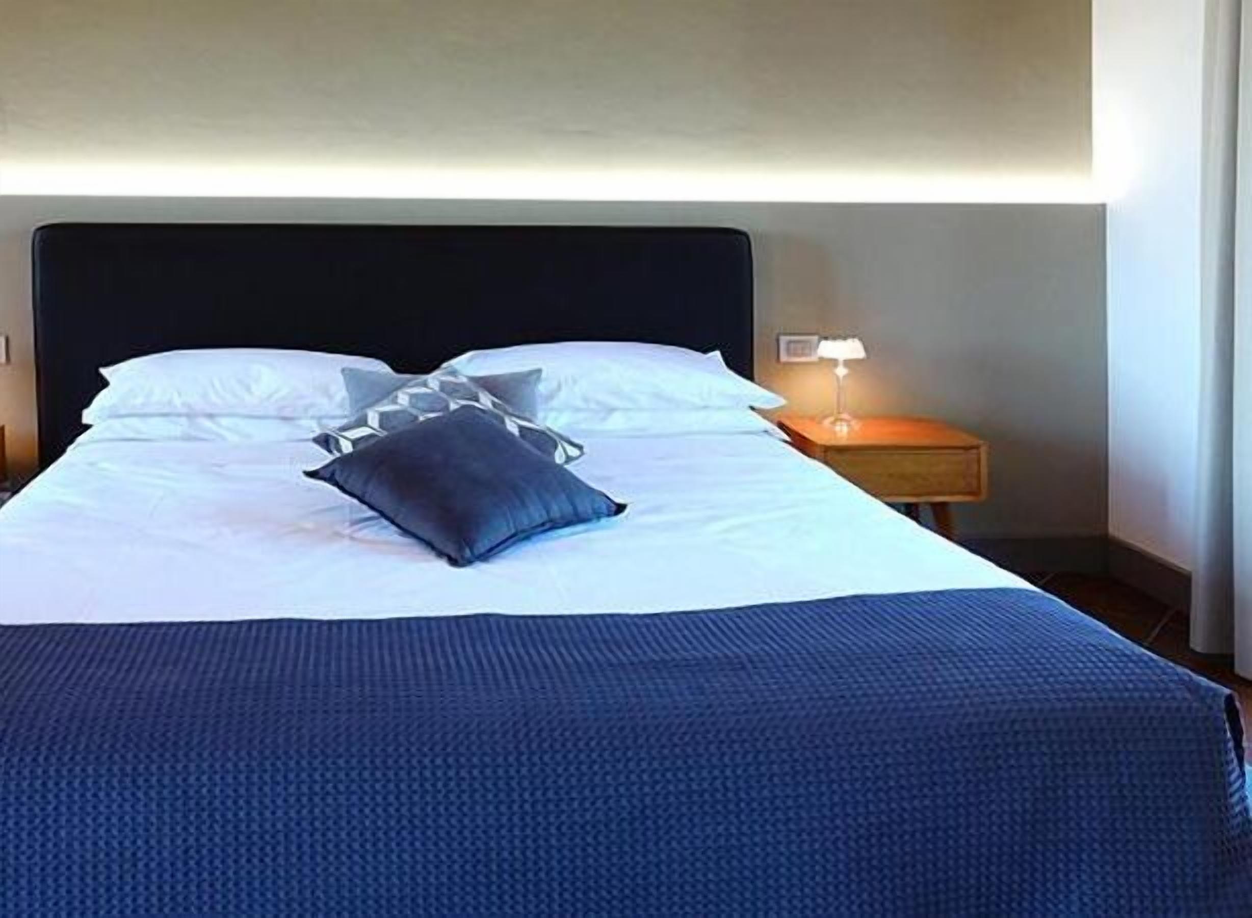 Ein ordentlich bezogenes Bett mit weißen Bettwäsche und einer dunkelblauen Decke, eine Nachttischlampe und ein Holznachttisch.