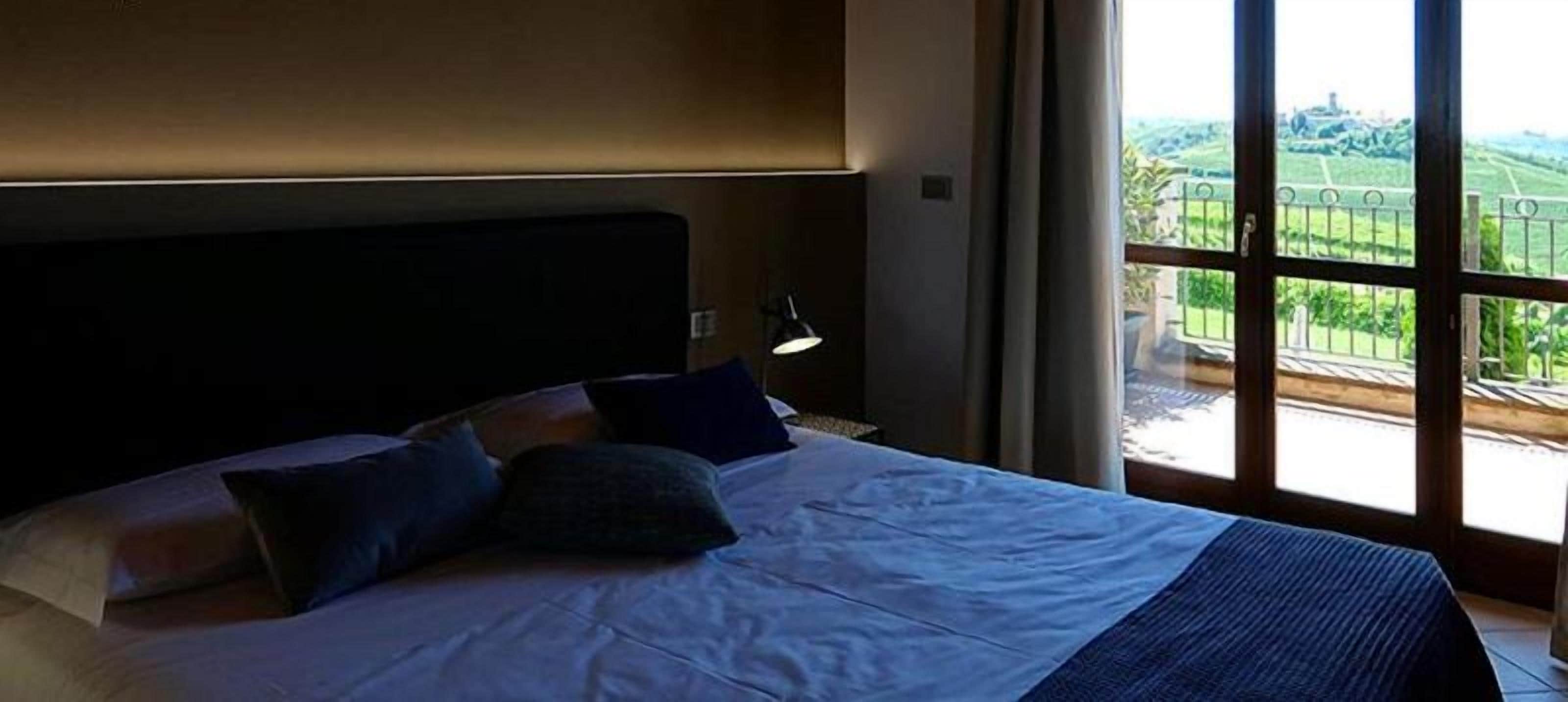A hotel room with a large bed, a nightstand, a lamp, and a view of the outdoors through a large window.