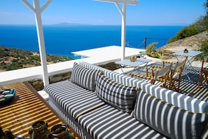A terrace with a sofa and chairs, overlooking a scenic view of the sea and hills. - Villa Alisachni (Andros)