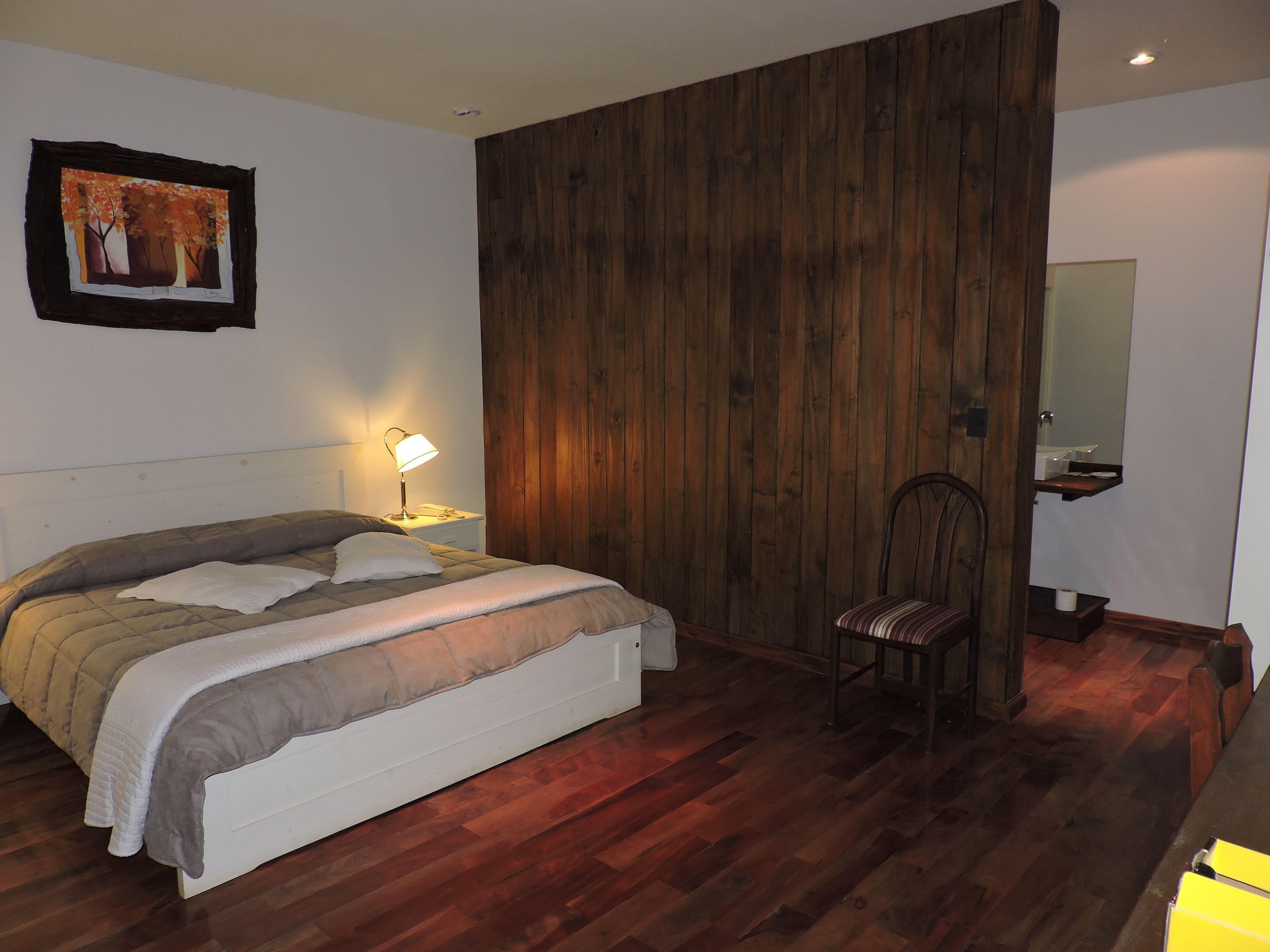 Quarto com piso de madeira, cama com colcha branca e cinza, mesa de cabeceira com abajur, uma cadeira e um painel divisor de madeira.