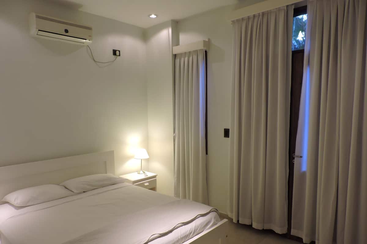 A bedroom with a bed, an air conditioning unit, a nightstand with a lamp, and a door with curtains.