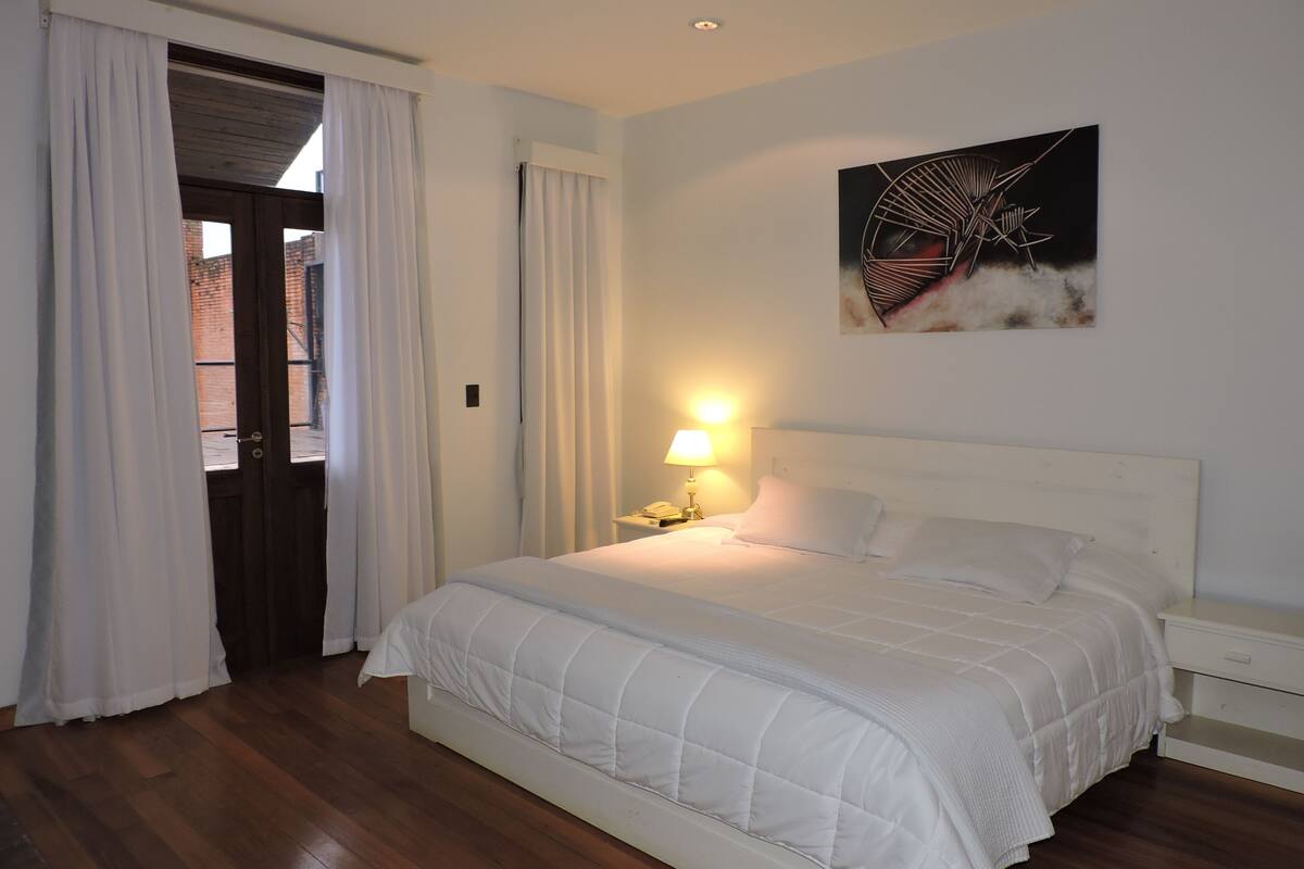A bedroom with a bed, white curtains, a nightstand with a lamp, and a painting on the wall.