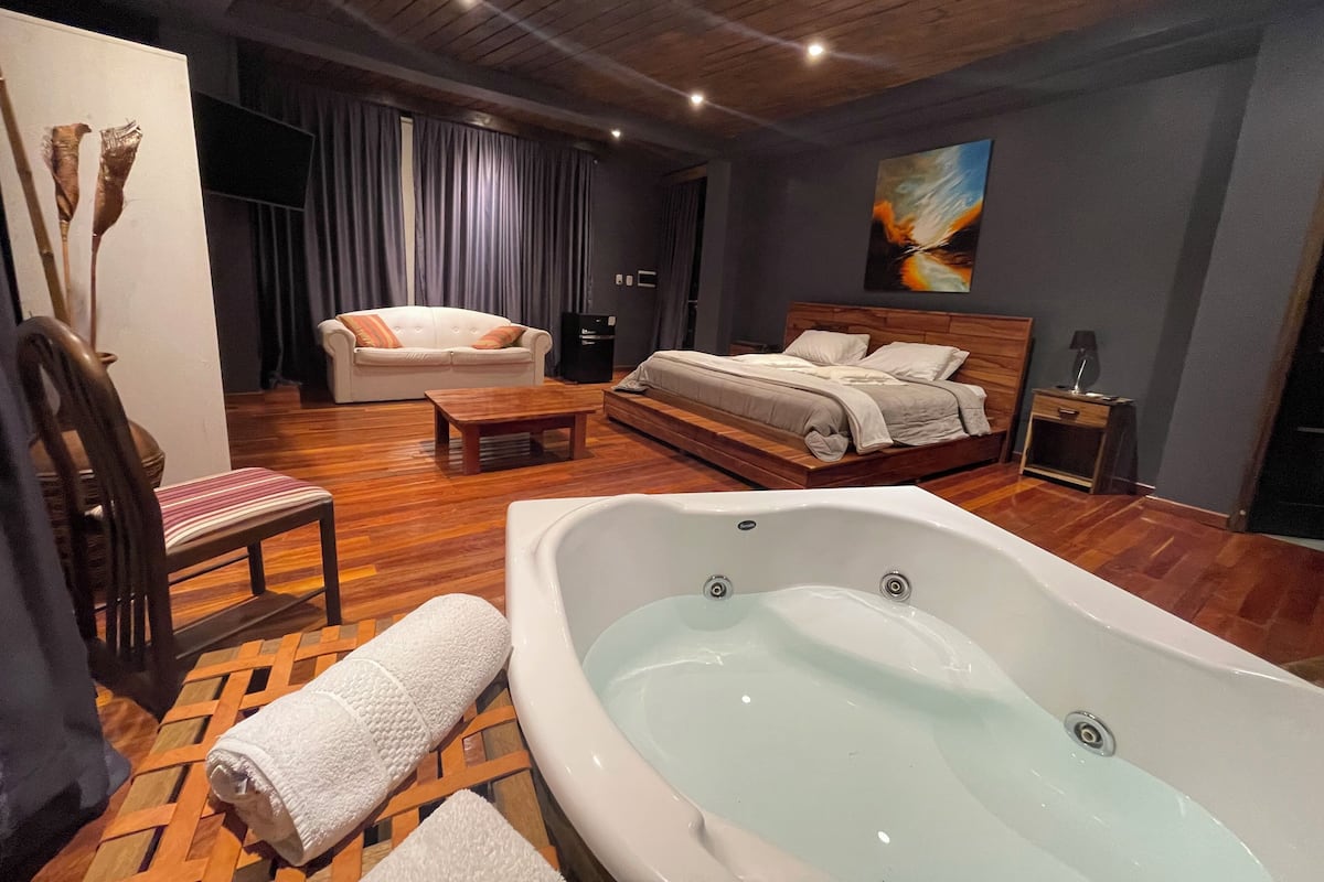 A modern bedroom with a large bathtub, a sofa, a wooden coffee table, and a bed with a headboard.
