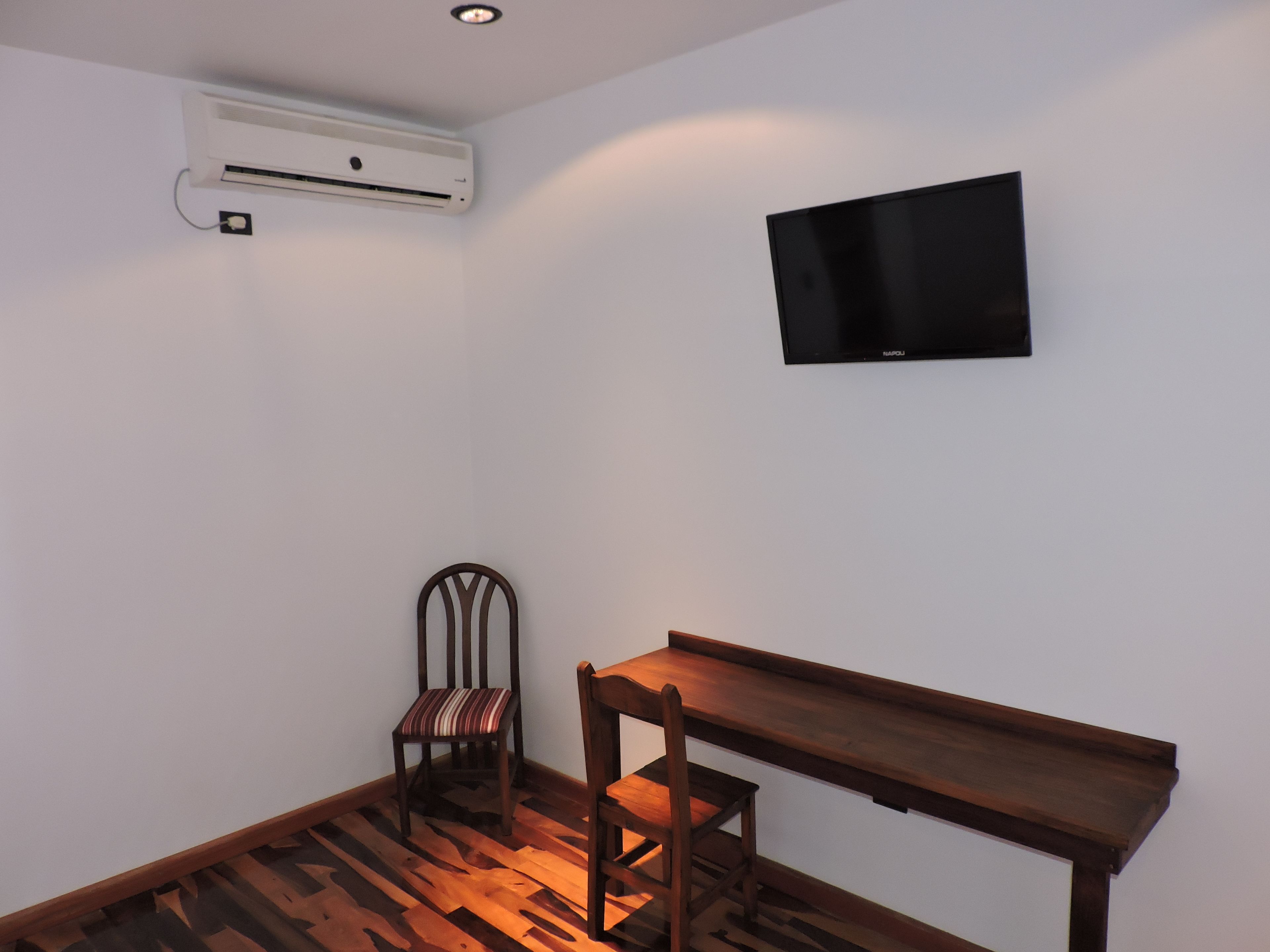A small room with a wooden floor, a single chair, a wooden table, a wall-mounted TV, and an air conditioning unit.