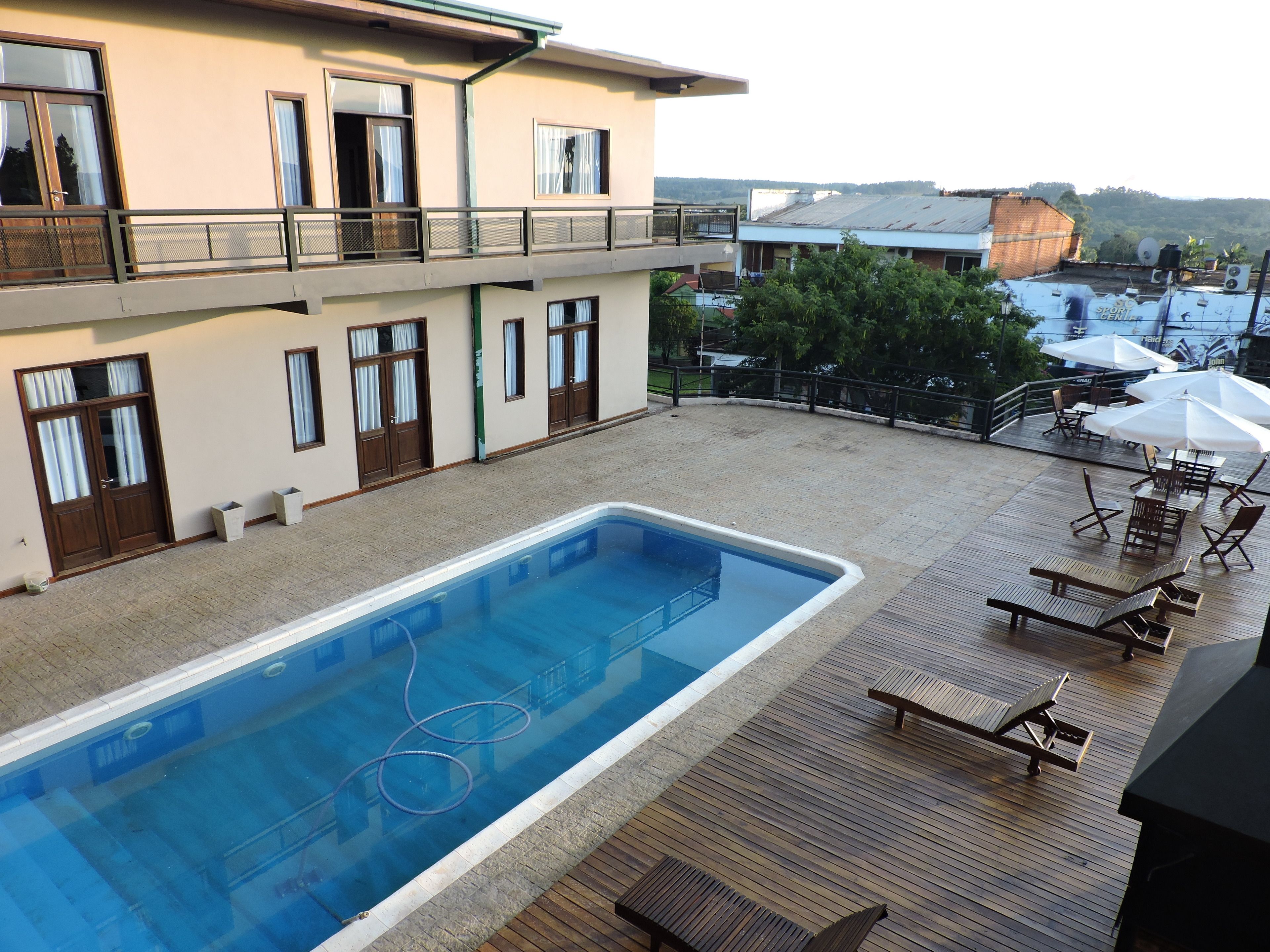A swimming pool with a wooden deck and lounge chairs, a building with multiple windows and balconies, and a view of the surrounding area.