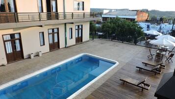 A swimming pool with a wooden deck and lounge chairs, a building with multiple windows and balconies, and a view of the surrounding area.