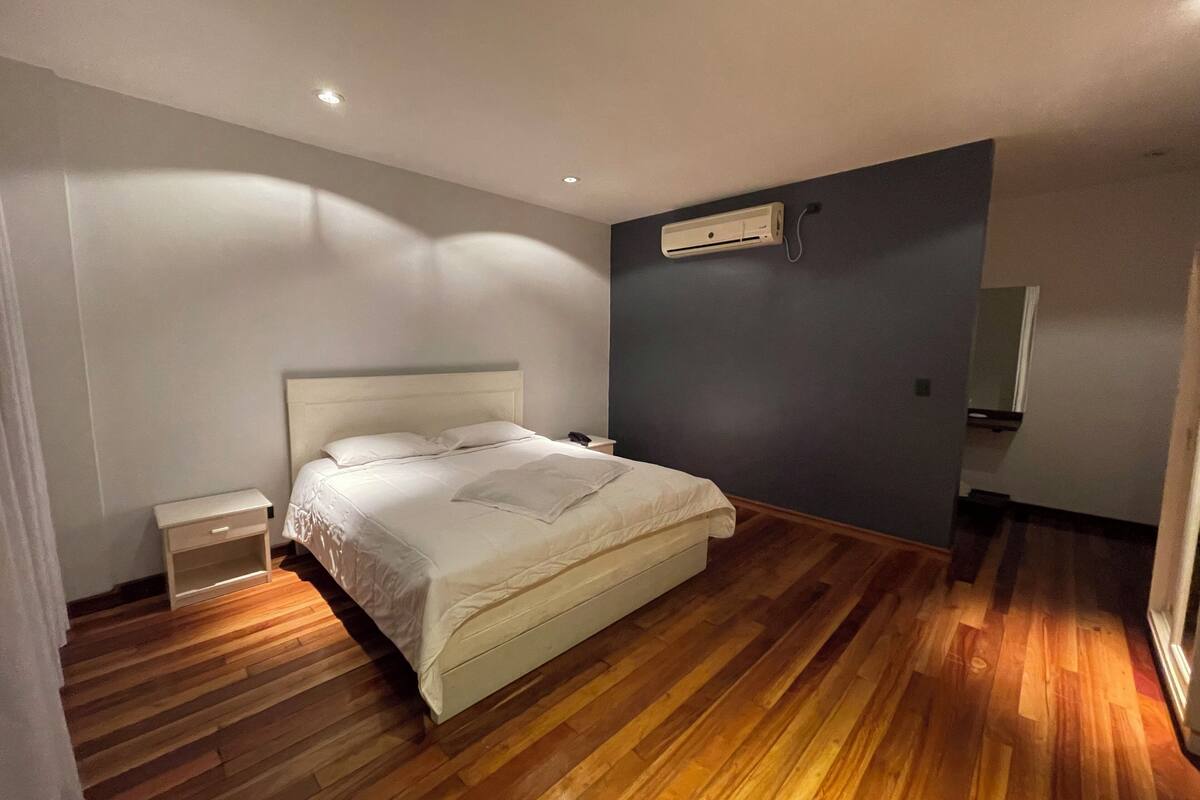 A bedroom with a bed, bedside tables, an air conditioning unit, and a wooden floor.