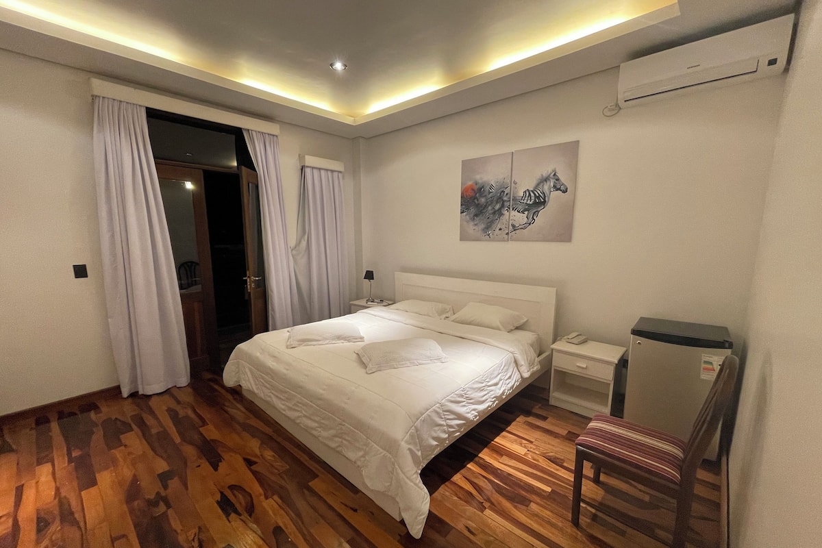A bedroom with a wooden floor, a bed with white linens, a nightstand, a chair, and a painting on the wall.