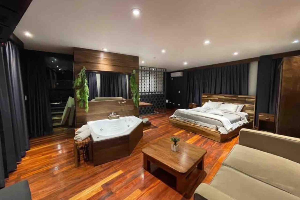 A modern hotel room with a large bed, a separate seating area with a sofa and a small table, a wooden floor, and a large window.