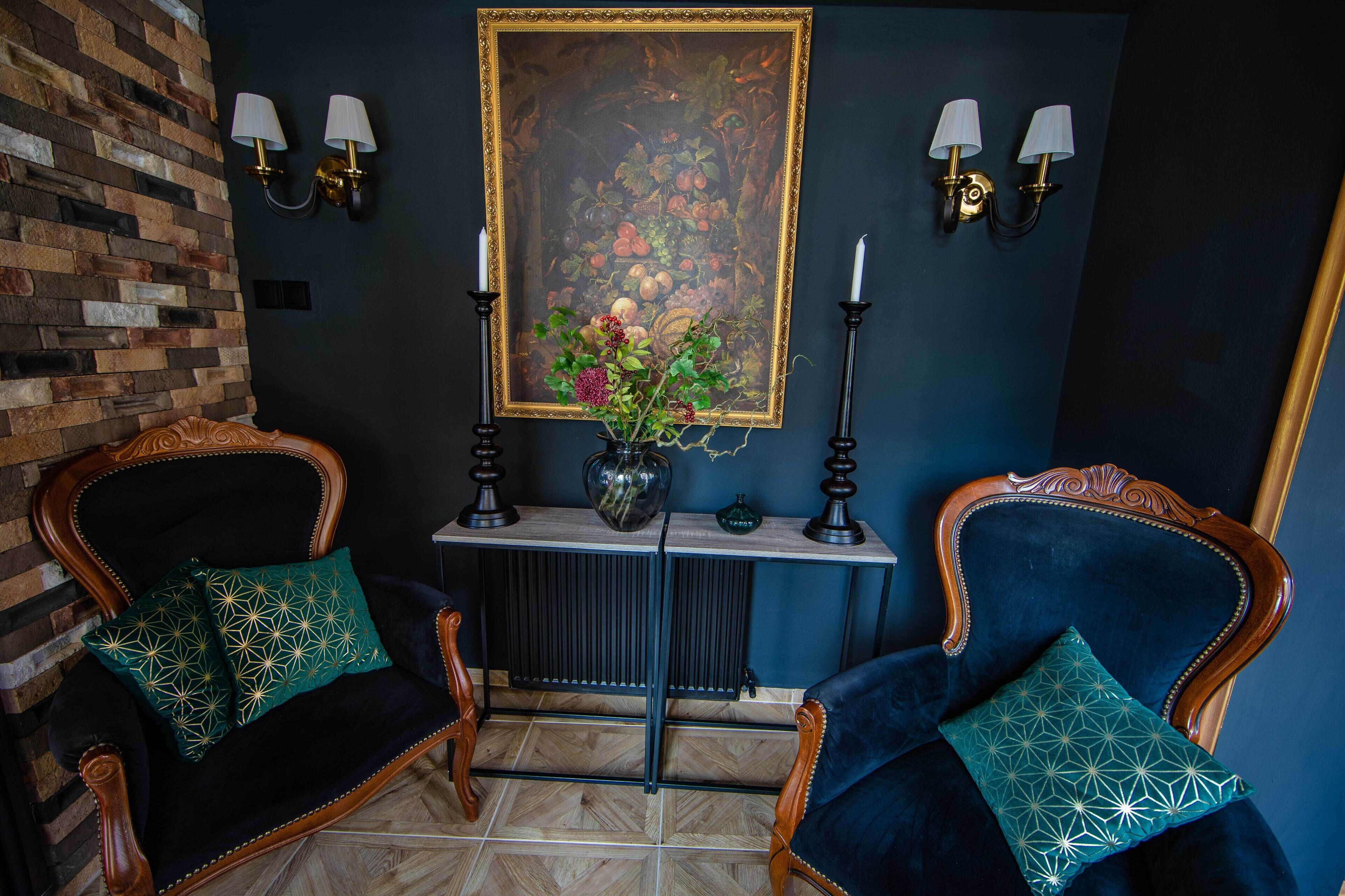 A room with two dark blue upholstered chairs with patterned cushions, a small table with a vase and a framed painting, and wall-mounted lamps.