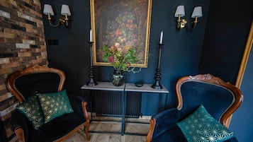 A room with two dark blue upholstered chairs with patterned cushions, a small table with a vase and a framed painting, and wall-mounted lamps.