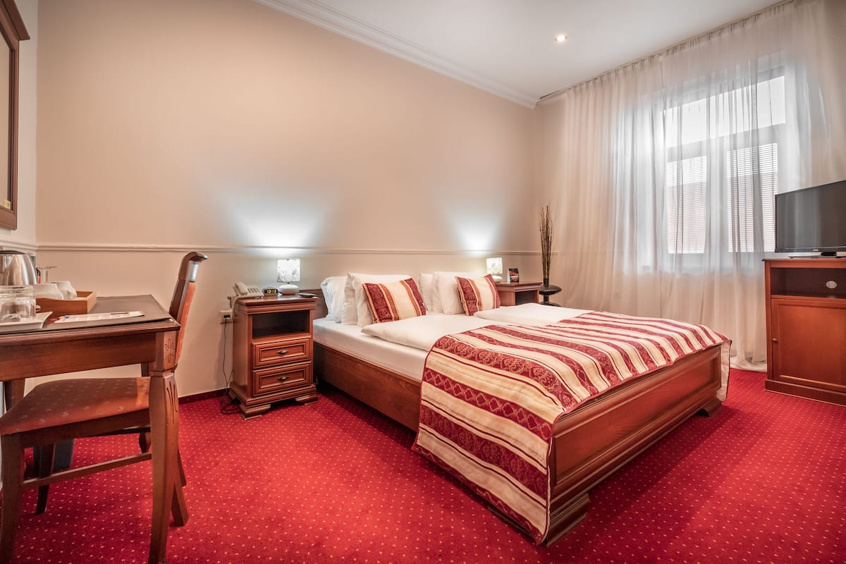 A hotel room with a large bed, a desk with a chair, a television, and a window with sheer curtains.