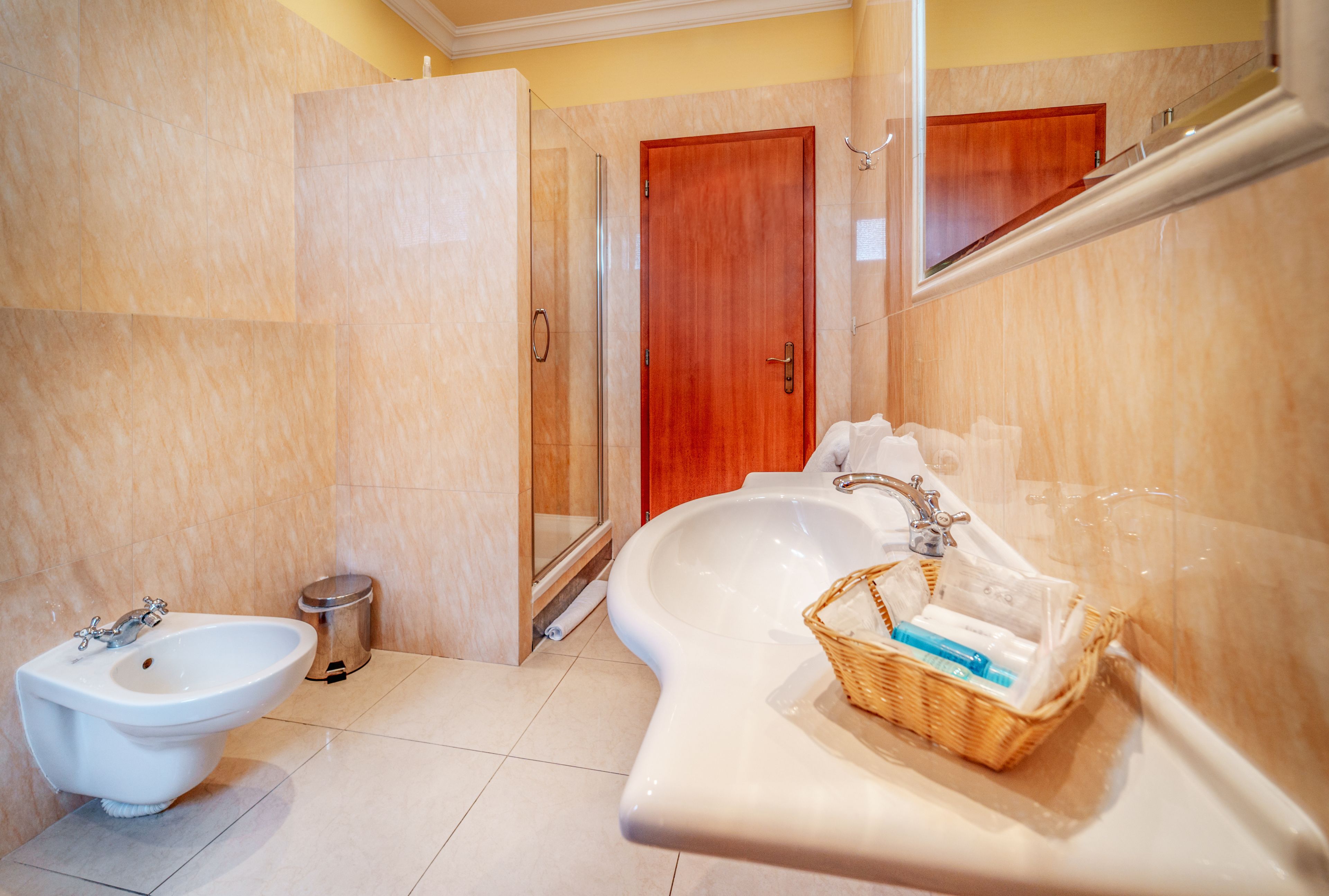 A bathroom with a bidet, a glass-enclosed shower, a wooden door, and a wicker basket on the counter.