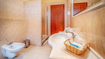 A bathroom with a bidet, a glass-enclosed shower, a wooden door, and a wicker basket on the counter.