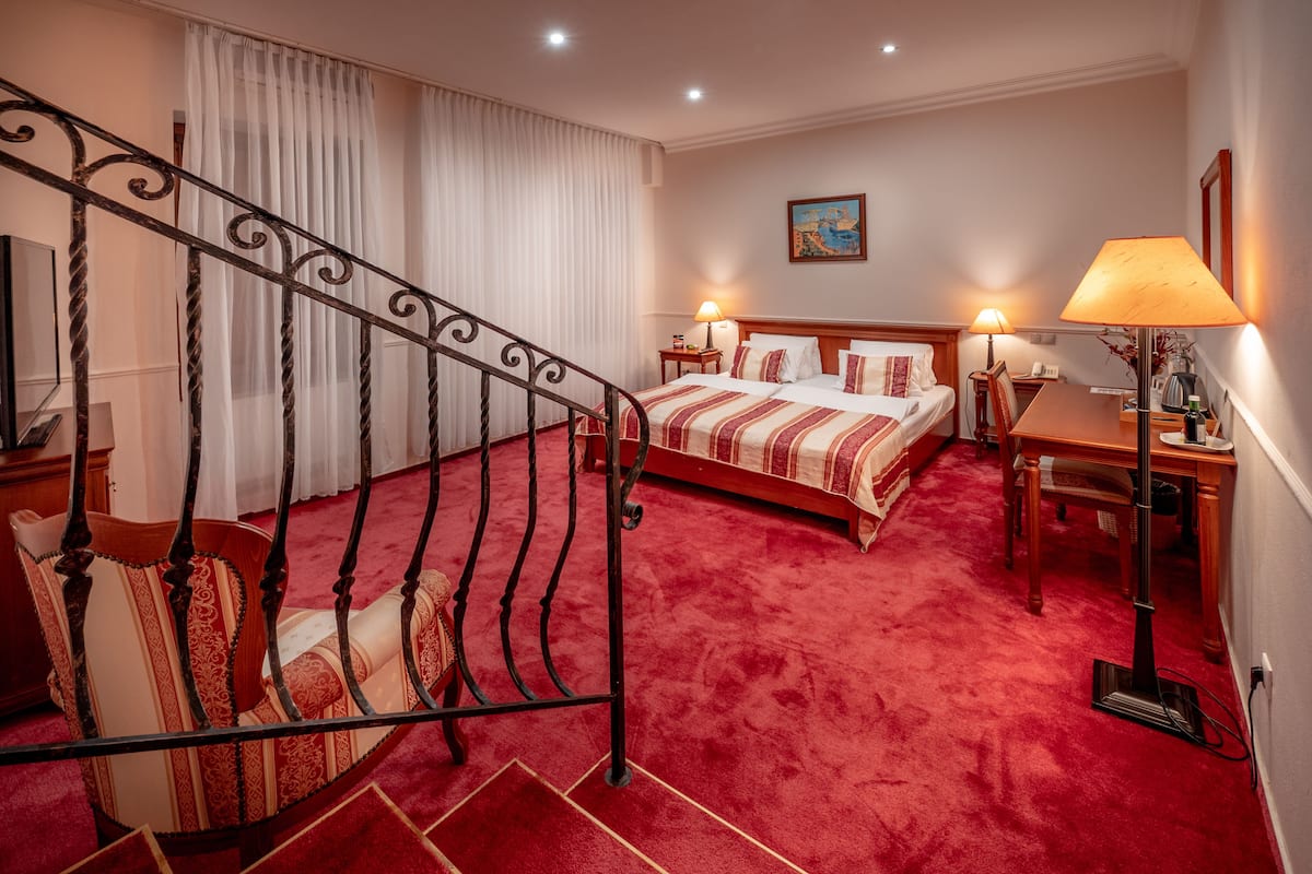 A hotel room with a red carpet, a bed with striped bedding, a desk with a lamp, and a painting on the wall.