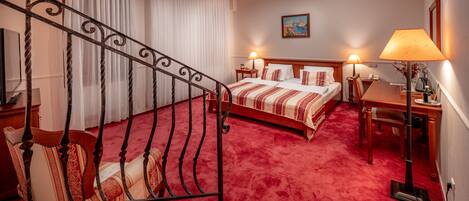 Una habitación de hotel con alfombra roja, cama con ropa de cama a rayas, escritorio con lámpara y un cuadro en la pared.