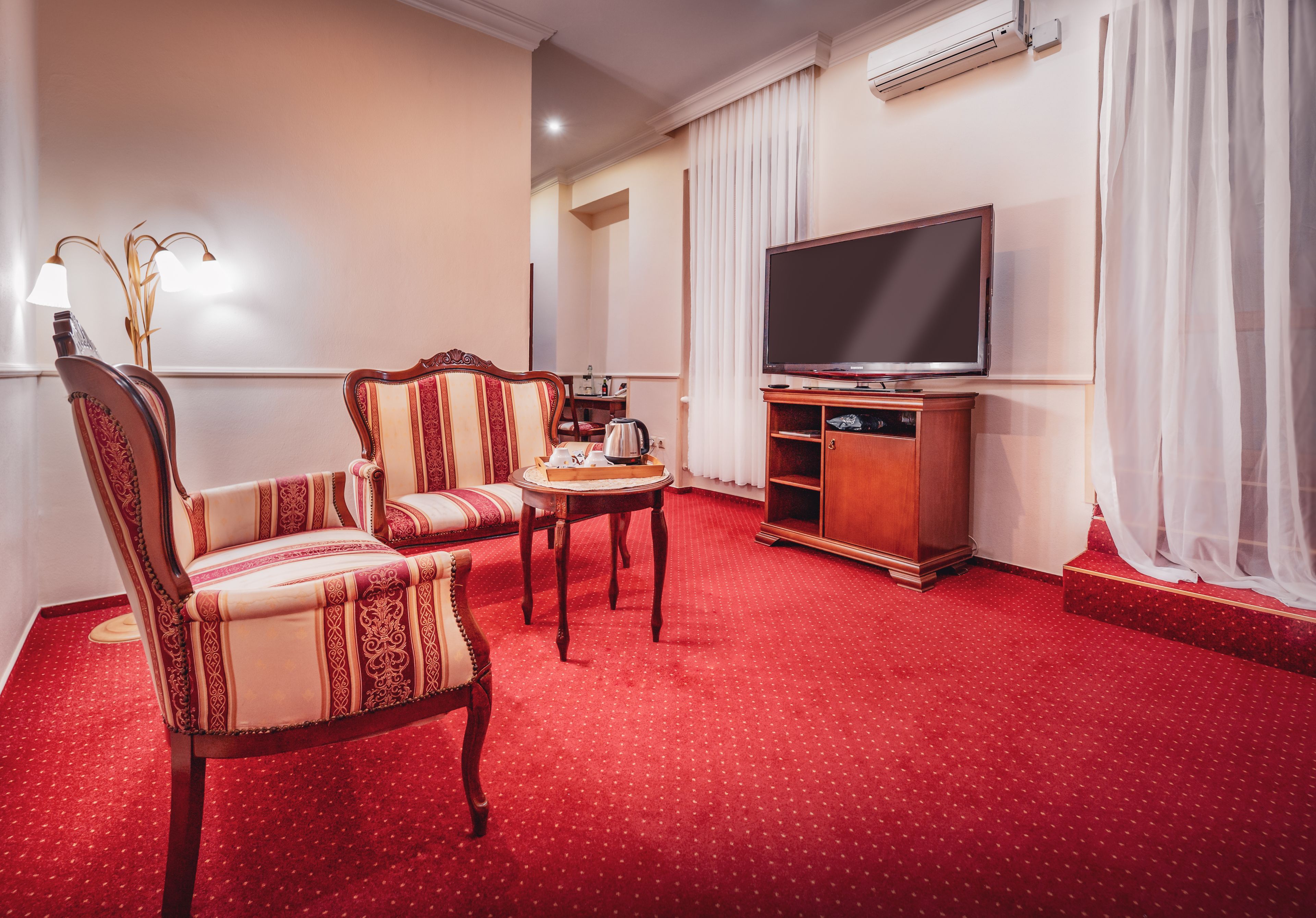 Habitació d'hotel amb una catifa vermella, un moble de televisió de fusta, un televisor de pantalla plana, una cadira de fusta i una taula petita amb un brollador.