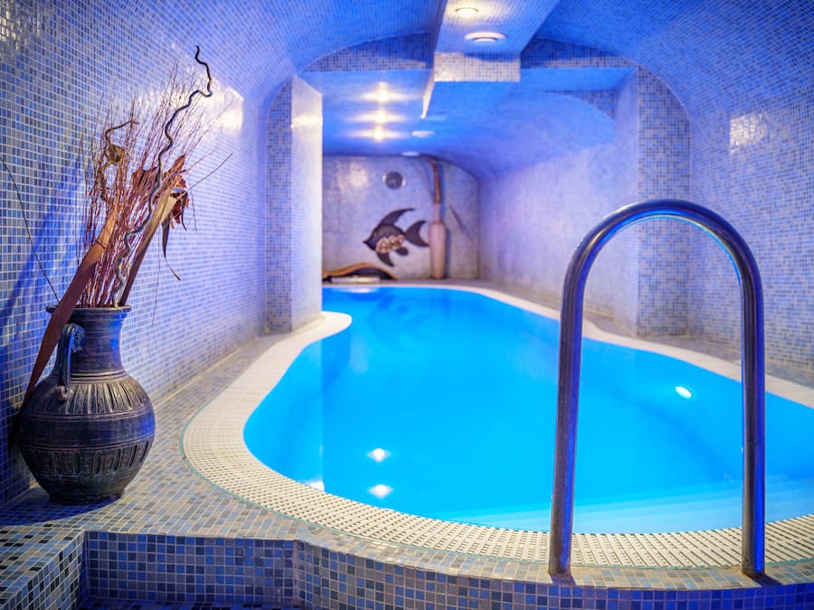 Una pequeña piscina interior con baldosas, una fuente de agua cristalina azul, un jarrón decorativo con ramas y un mural de un pez en la pared.