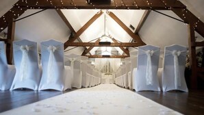 Un pasillo nupcial con sillas blancas y una alfombra blanca, bajo una estructura de madera con luces colgantes.