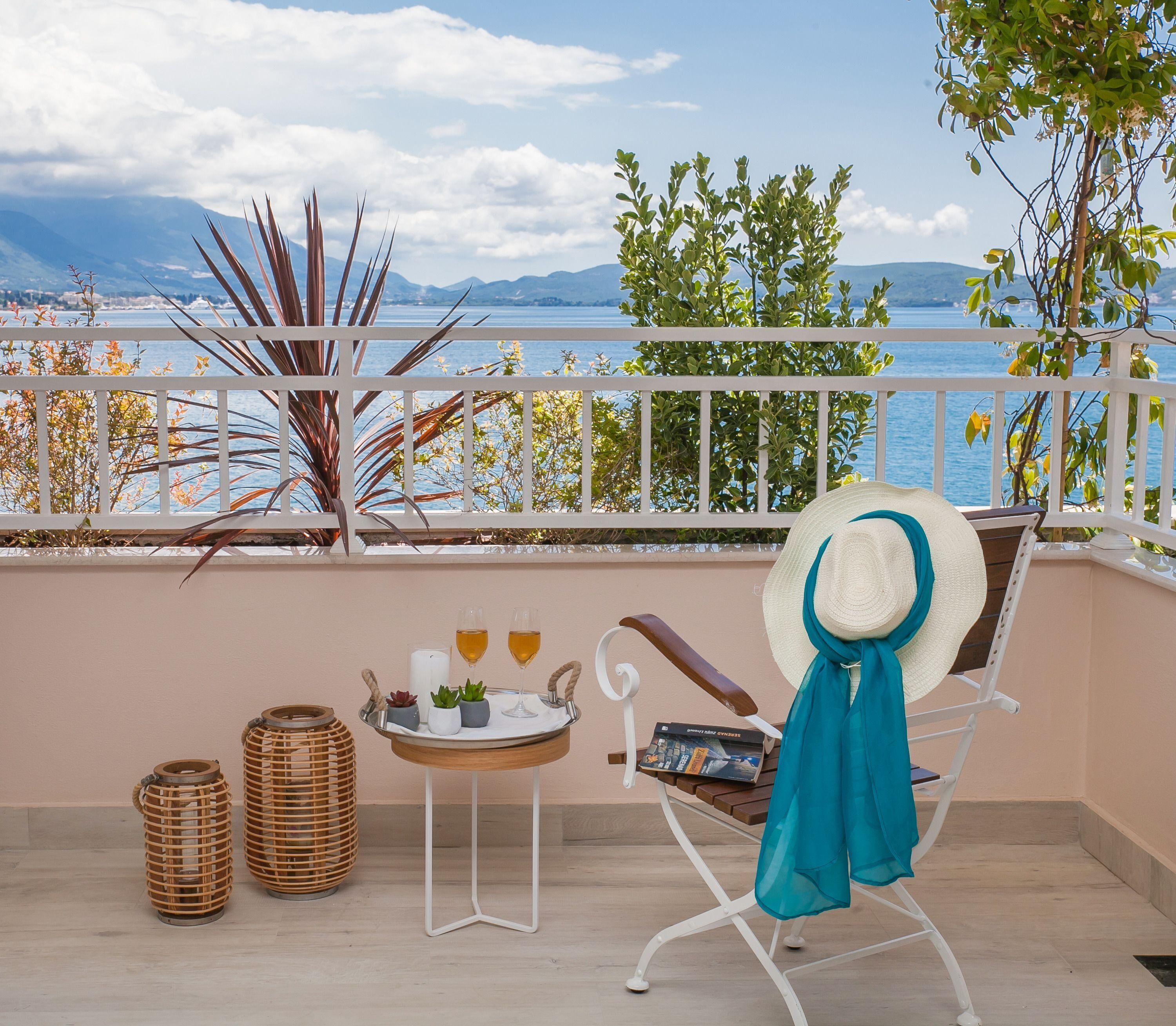 Un balcon avec vue sur la mer, une chaise avec un chapeau, un plateau avec des boissons et un livre, et des paniers en osier.