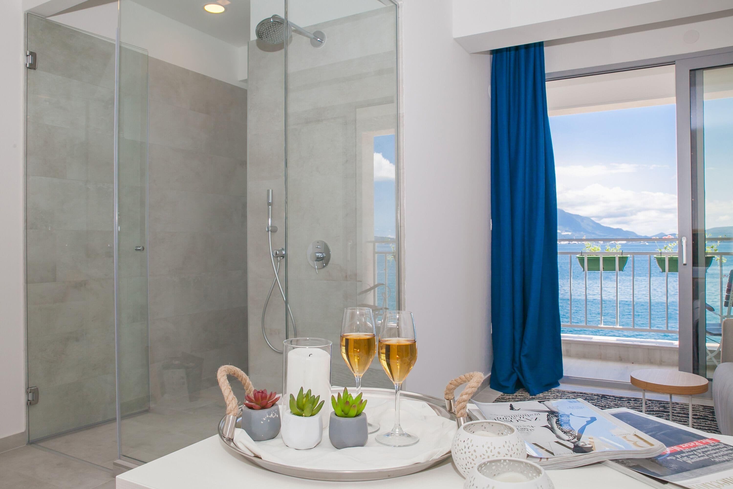 Ein modernes Badezimmer mit Duschkabine aus Glas, einem großen Fenster mit Meerblick sowie einem Tablett mit zwei Sektgläsern und kleinen Pflanzen.