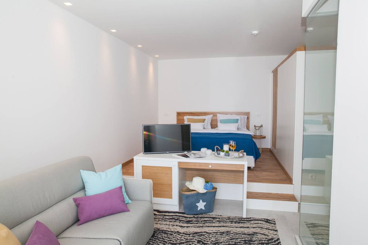A modern bedroom with a bed, a desk with a laptop, a TV, and a glass shower area.