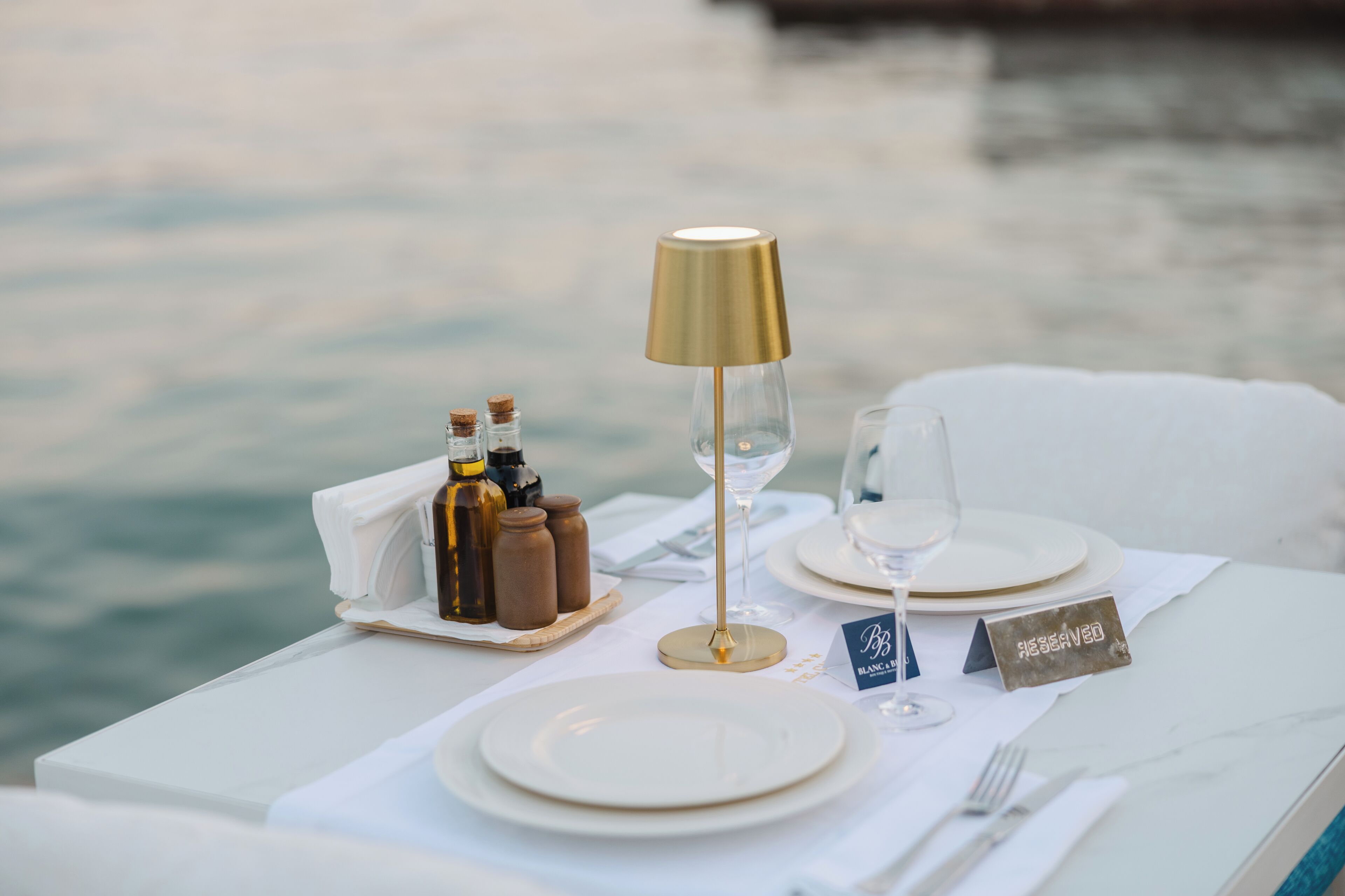Mesa preparada para comer con vista al agua, una lámpara, saleros y pimenteros, y una copa de vino.